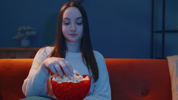 Nő ül a kanapén egyedül, eszik popcorn és érdekes TV sorozat, horror film otthon — Stock videók