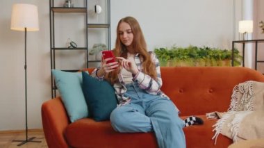 Kanepede oturan neşeli genç kadın, sosyal medya uygulamasında cep telefonu paylaşımı mesajları kullanıyor.