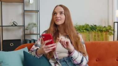 Kanepede oturan neşeli, genç bir kadın akıllı telefon kullanıyor video izliyor, online alışveriş yapıyor, dinliyormuş.