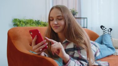 Neşeli genç bir kadın kanepeye uzanıp akıllı telefon kullanıyor video izliyor, online alışveriş yapıyor, telefon çekiyor.