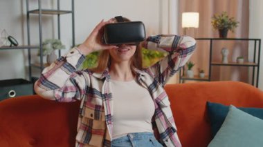 Genç bir kadın evde sanal gerçeklik kulaklığı kullanıyor, video konseptinden zevk alıyor, ellerini havada hareket ettiriyor.