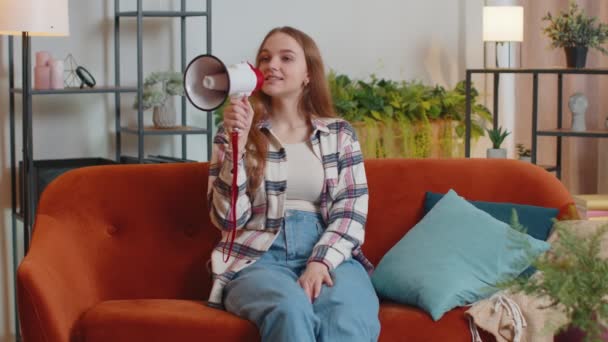 Молода жінка кричить в мегафоні гучномовець оголошує знижки на продаж нерухомості вдома на дивані — стокове відео