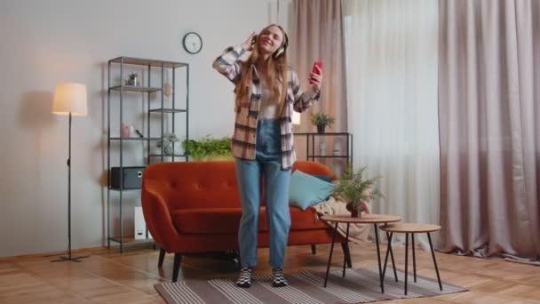 Mujer joven muy alegre en auriculares inalámbricos bailando, cantando en un sofá acogedor en la sala de estar en casa — Vídeos de Stock