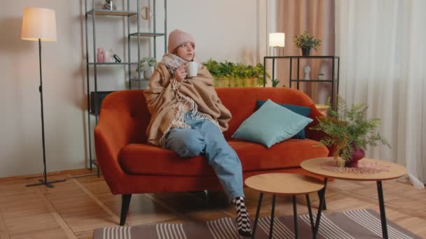 年轻的生病妇女戴着裹着格子花的帽子，一个人坐在沙发上，冷得发抖，喝着热茶 — 图库视频影像