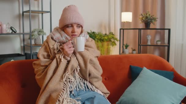 Νεαρή άρρωστη γυναίκα φοράει καπέλο τυλιγμένο σε καρό κάθεται μόνη της τρέμοντας από το κρύο στον καναπέ πίνοντας ζεστό τσάι — Αρχείο Βίντεο