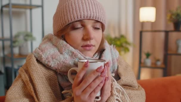 Jonge zieke vrouw draagt hoed gewikkeld in geruite zitten alleen rillen van de kou op de bank het drinken van warme thee — Stockvideo