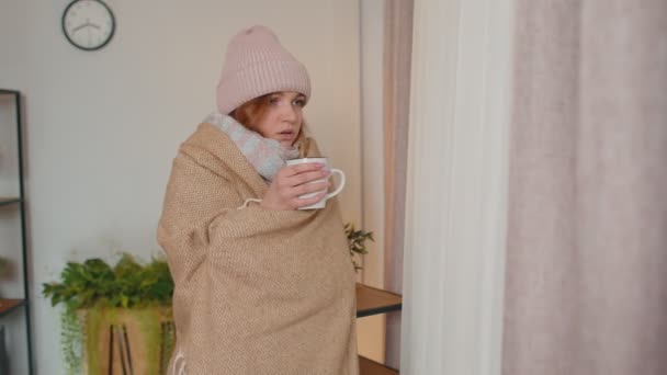 Młoda chora kobieta nosi kapelusz zawinięty w kratę. Trzęsie się z zimna na kanapie. Pije gorącą herbatę. — Wideo stockowe