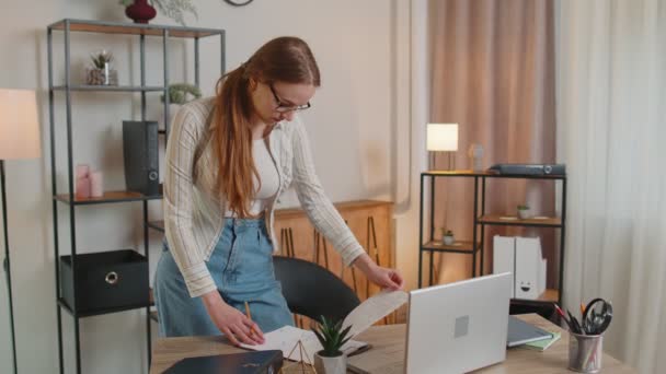 Menina jovem de pé por escrivaninha em casa escritório, trabalhando em documentos de papel analisa dados cheques — Vídeo de Stock