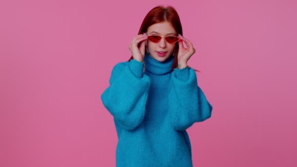 Seductive cheerful redhead girl in blue sweater wearing sunglasses, charming smile on pink wall — Stock Video