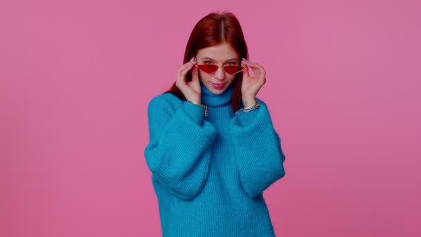 Sincero legal alegre ruiva menina em suéter azul usando óculos de sol, sorriso encantador na parede rosa — Vídeo de Stock