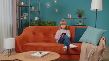 Küçük ilkokul çocuğu cep telefonu teknolojisi kullanıyor evde tek başına kanepede oturuyor.