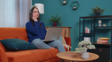 Kanepede oturan neşeli, genç esmer kadın ev telefonuyla konuşan dizüstü bilgisayar kullanıyor.