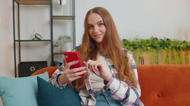 Kanepede oturan neşeli, genç bir kadın akıllı telefon kullanıyor video izliyor, online alışveriş yapıyor, dinliyormuş.