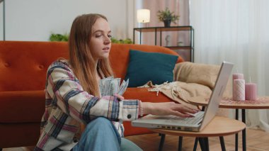 Gülümseyen genç bir kadın nakit para sayıyor ve bilgisayarını kullanıyor. Ev faturalarını hesaplıyor.