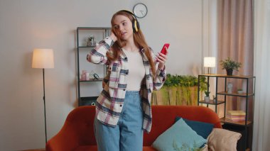 Kulaklık takan, müzik dinleyen, evdeki oturma odasında şarkı söyleyen beyaz bir kadın.