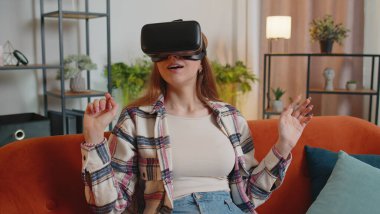 Genç kız evde sanal gerçeklik kulaklığı kullanıyor. 3 boyutlu video oyunu oynuyor. Ellerle jestler yapıyor.