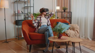 Genç bir kadın evde sanal gerçeklik kulaklığı kullanıyor, video konseptinden zevk alıyor, ellerini havada hareket ettiriyor.