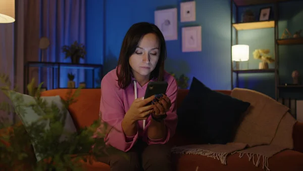 Kobieta SMS-uje na smartfonie, przewija się na telefonie komórkowym i siedzi sama na kanapie w domu — Zdjęcie stockowe