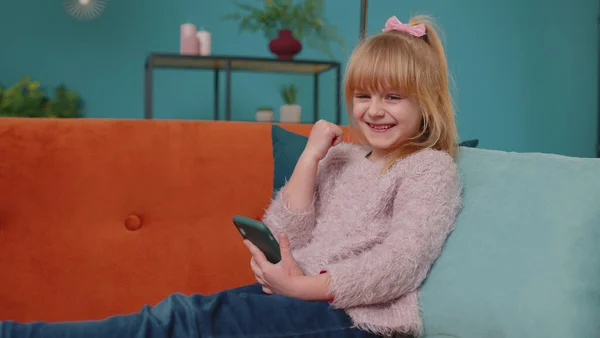 Glad liten skola unge flicka använda mobiltelefon gör vinnaren gest fira ensam hemma soffa — Stockfoto