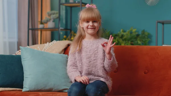 Portret van een klein kind meisje kind alleen op de bank thuis met ok gebaar, als teken, positief goed — Stockfoto