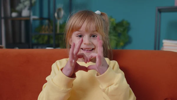 Usmívající se dítě dívka dítě na domácí pohovce při pohledu na kameru dělá srdce gesto demonstruje znamení lásky — Stock fotografie