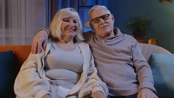 Счастливая пожилая пара пожилых людей обнимает, смеется, улыбается, глядя в камеру на домашнем диване — стоковое фото