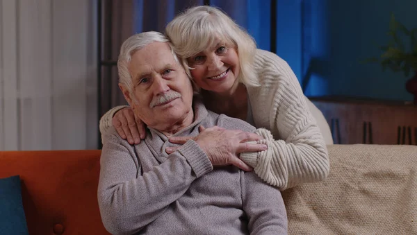 Happy old senior elderly family couple hugging, smiling, smiling looking at camera at home sofa