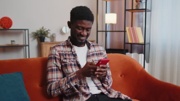 Χαρούμενος νεαρός ενήλικας κάθεται στον καναπέ χρησιμοποιώντας το smartphone βλέποντας βίντεο, online αγορές, χτυπώντας — Αρχείο Βίντεο