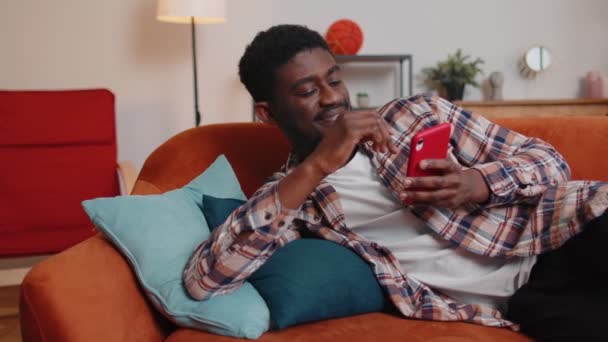 Веселый молодой человек лежит на диване, обмениваясь сообщениями в социальных сетях — стоковое видео