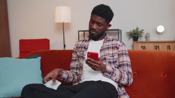 Szomorú afro-amerikai férfi kezében készpénz és a mobiltelefon kiszámítja hazai számlák otthon — Stock videók