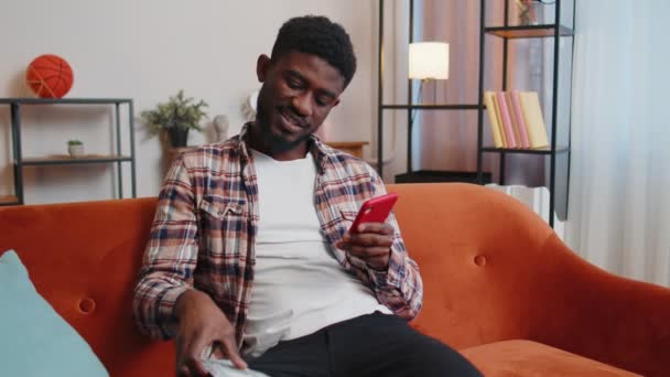 Усміхнений щасливий молодий чоловік рахує гроші готівкою і використовує мобільний телефон для розрахунку внутрішніх рахунків вдома — стокове відео
