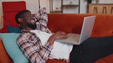 Genç adam dizüstü bilgisayar kullanıyor. Evdeki kanepede yatıyor. Çalışıyor, online alışveriş yapıyor, film izliyor.
