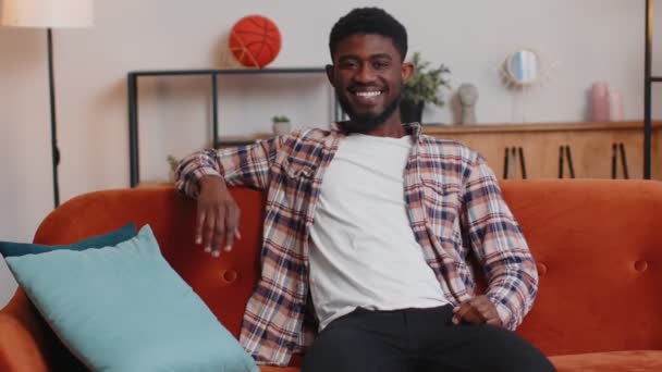 Mladý muž se přátelsky usmívá před kamerou, mává rukama a mává na pozdrav, zdravím, zdravím vás doma na pohovce — Stock video