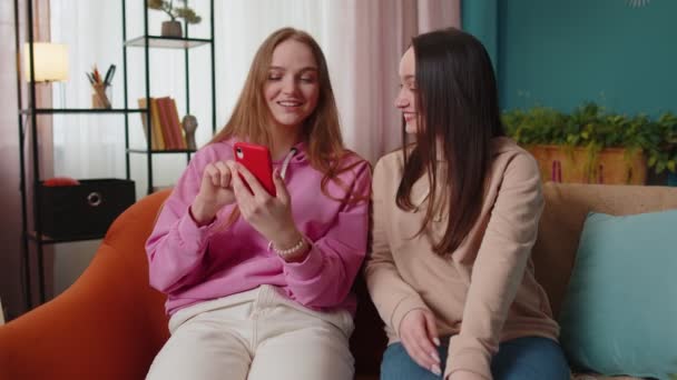 Веселые девушки друзья братья и сестры смотрят фотографии на смартфоне на диване дома, пользуются социальными сетями — стоковое видео