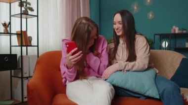 Neşeli kız arkadaşlar kardeşler evdeki kanepede akıllı telefondan fotoğraf izleyip sosyal medyanın tadını çıkarıyorlar.