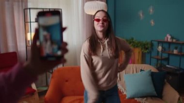 Neşeli blogcu genç kız evde cep telefonuyla video çekerek dans ediyor.