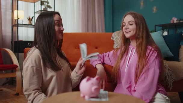 Freundinnen Geschwister sitzen auf dem Boden und werfen abwechselnd Dollarnoten in Sparschweine — Stockvideo
