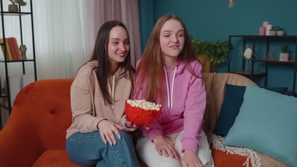 Dos chicas amigos hermanos viendo la película favorita de la serie de televisión, comiendo palomitas de maíz, sentado en el sofá en casa — Vídeos de Stock