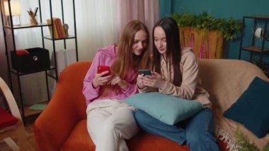 Neşeli kız arkadaşlar kardeşler evdeki kanepede akıllı telefondan fotoğraf izleyip sosyal medyanın tadını çıkarıyorlar.