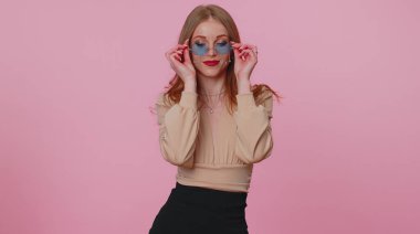 Baştan çıkarıcı esmer, bej bluzlu, güneş gözlüklü, pembe duvarda büyüleyici bir gülümsemesi olan kız.
