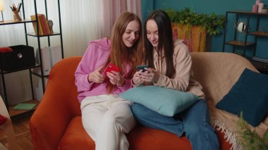 Neşeli kız arkadaşlar kardeşler evdeki kanepede akıllı telefondan fotoğraf izleyip sosyal medyanın tadını çıkarıyorlar.