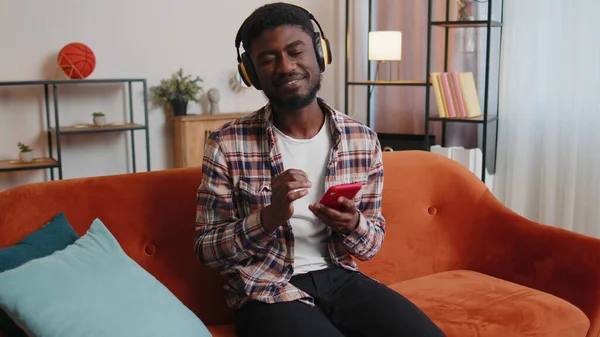 Kablosuz kulaklıklı neşeli genç adam dans ediyor, evdeki oturma odasında rahat koltukta şarkı söylüyor.