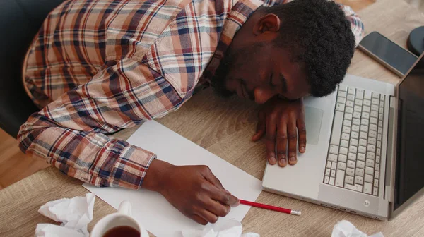 Zmęczony mężczyzna w biurze zasypiający na stole z laptopem, pogniecione kartki papieru — Zdjęcie stockowe