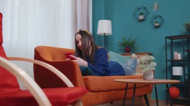 Rahatlamış, gülümseyen beyaz bir kadın. Kanepede uzanmış, akıllı telefonları kaydırıyor, internette geziniyor.