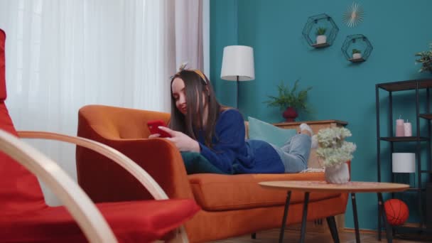 Relaks uśmiechnięta młoda kobieta kaukaska leżąca na domowej kanapie ze smartfonem przewijanie, przeglądanie online — Wideo stockowe
