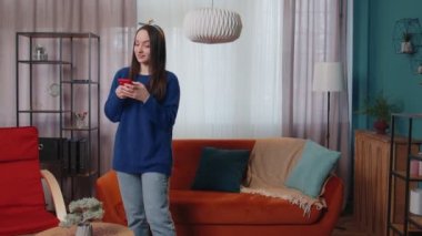 Rahatlamış, gülümseyen beyaz bir kadın akıllı telefonuyla evde duruyor, internette geziniyor.