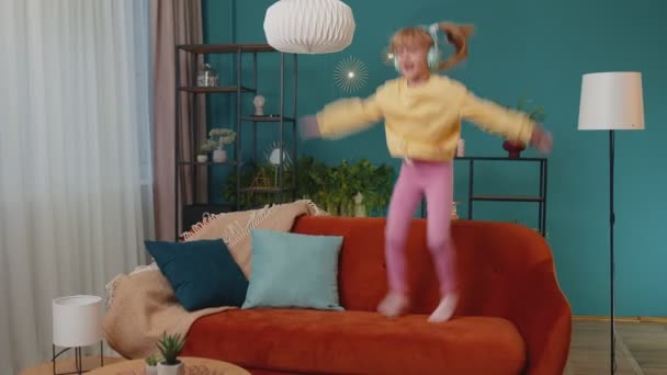 Щаслива дитина дівчинка в навушниках танцює і стрибає, слухаючи музику вдома наодинці — стокове відео