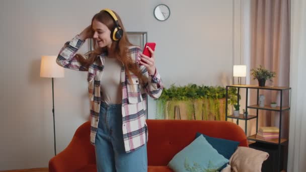 Överlycklig ung kvinna i trådlösa hörlurar dansar, sjunger på mysig soffa i vardagsrummet hemma — Stockvideo