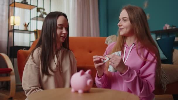 Lány barátok testvérek ül a padlón, és felváltva dobja dollár bankjegy malac bank — Stock videók
