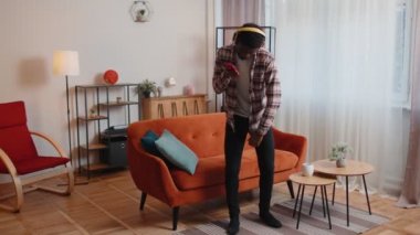 Kablosuz kulaklıklı neşeli genç adam dans ediyor, evdeki oturma odasında rahat koltukta şarkı söylüyor.